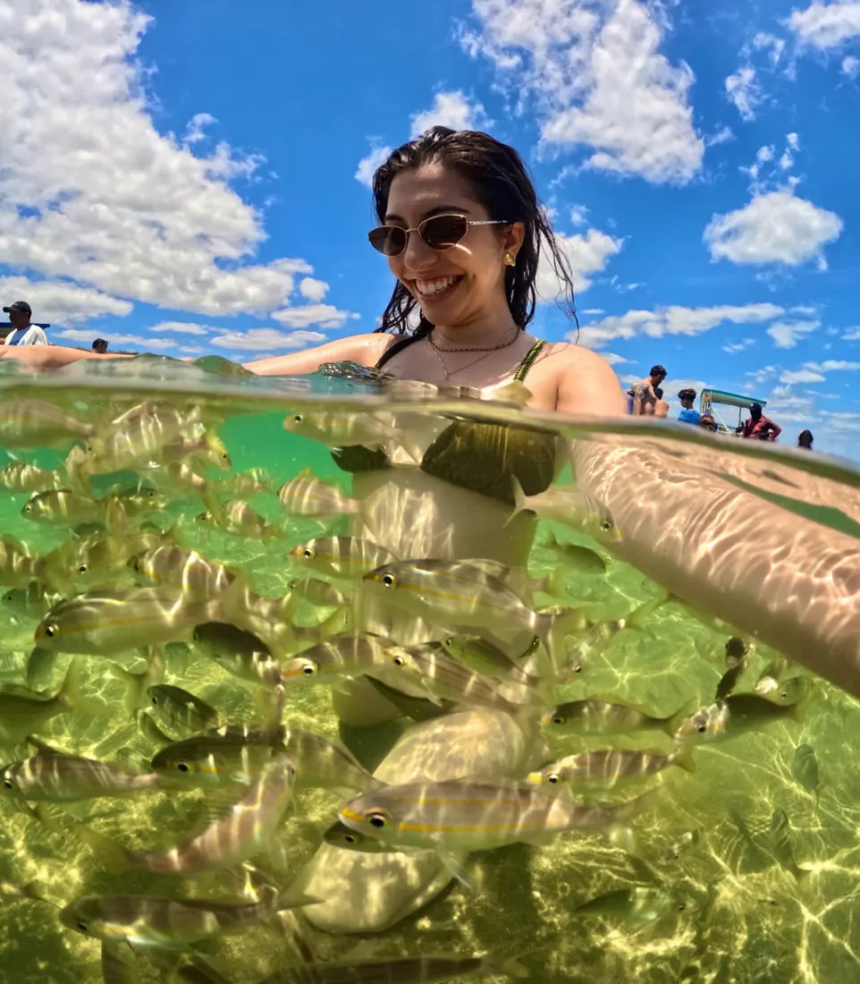Turista na piscina natural, metade dentro da água, com um cardume de peixes